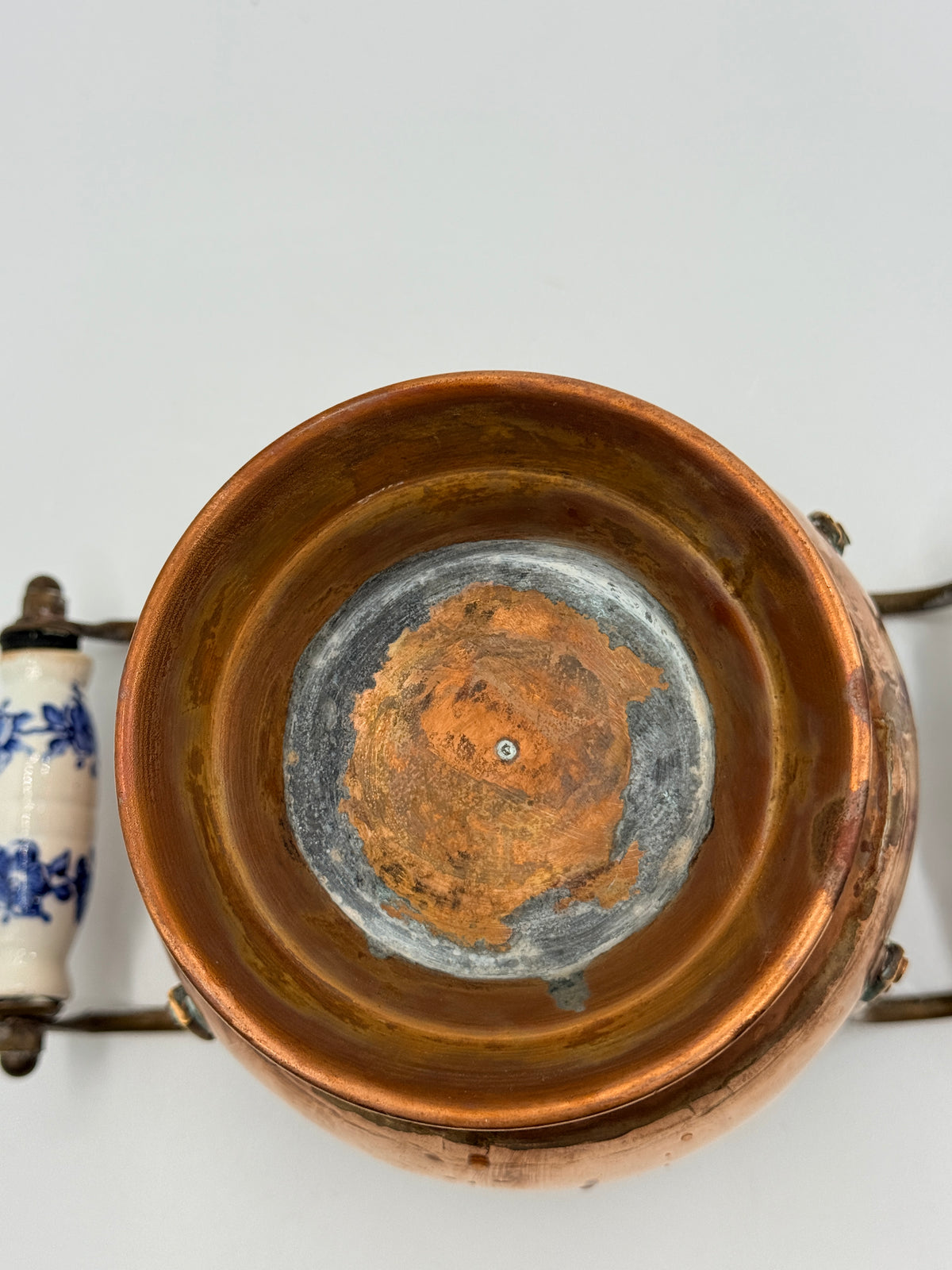 An antique copper pot shown from a top-down view, featuring a rustic orange-brown patina inside and a blue-and-white ceramic handle on the left, set against a plain light gray background.