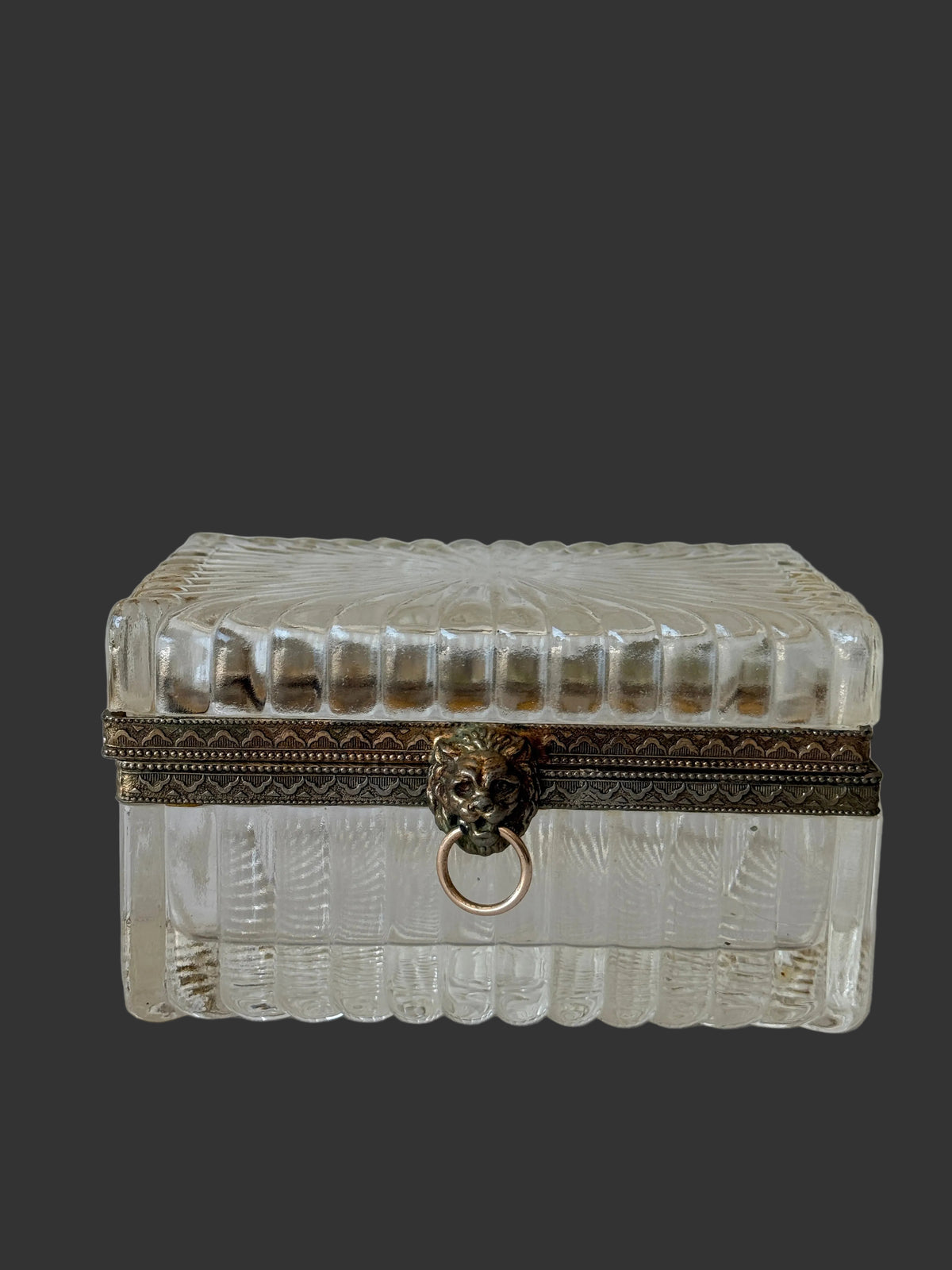 A vintage clear glass jewelry box with a textured, ribbed pattern, a dark brown decorative band, and an antique bronze lion-head ring clasp, set against a white paneled background.
