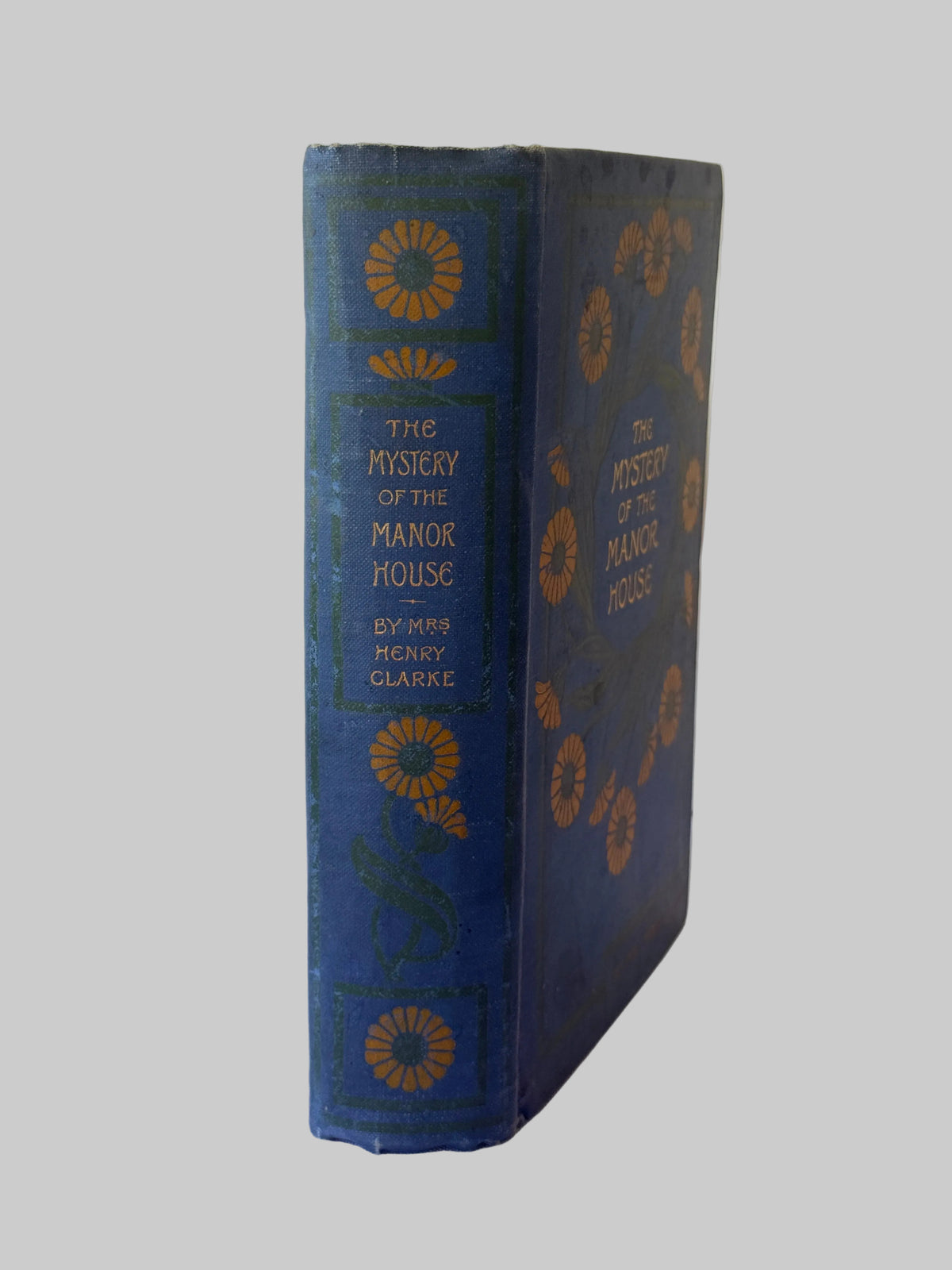 A vintage hardcover book titled 'The Mystery of the Manor House' by Henry Clarence, featuring a dark blue cover with gold floral patterns, photographed on a white paneled surface.