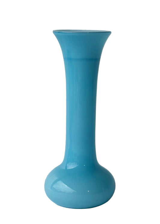 A tall, slender turquoise blue glass vase with a flared rim and a rounded base, standing on a light-colored surface against a white paneled background.