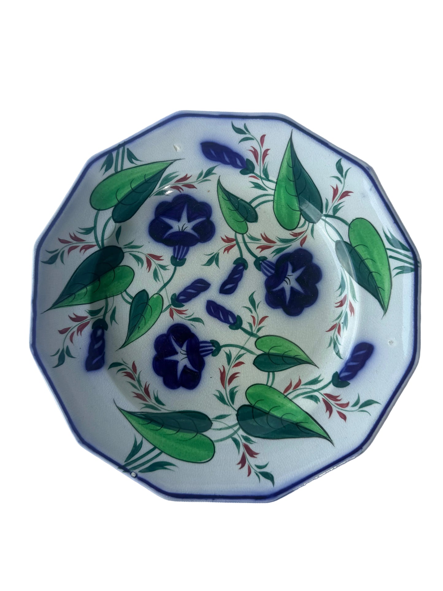 A round ceramic plate with a hand-painted floral pattern featuring dark blue flowers, green leaves, and small red accents on a white background, photographed from above on a light surface.