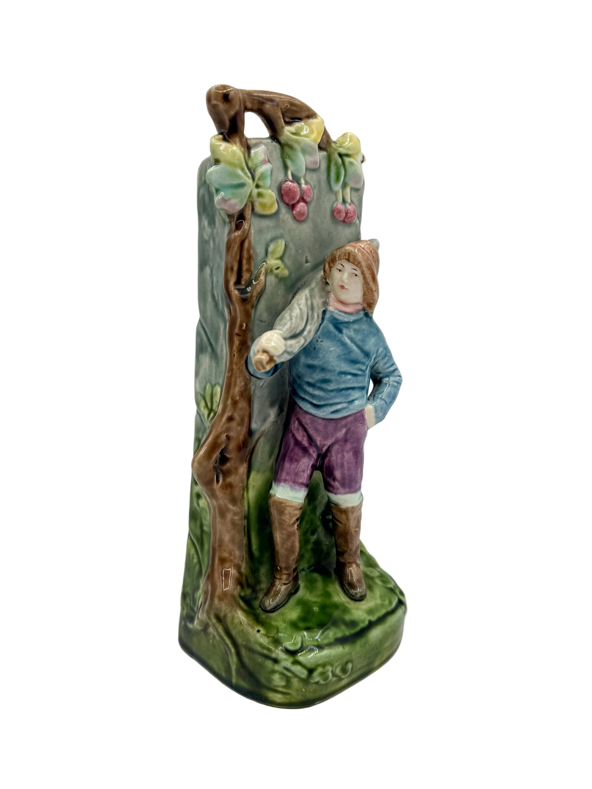 A vintage ceramic figurine of a young boy standing in front of a tree with cherries, glazed in earthy green, brown, blue, and purple tones.