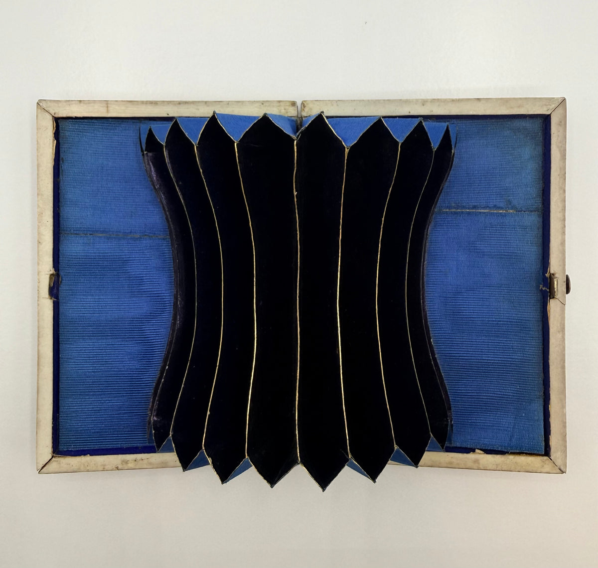 A square clap with a blue fabric-lined interior and a central accordion-style black pleated panel, photographed against a plain off-white wall.