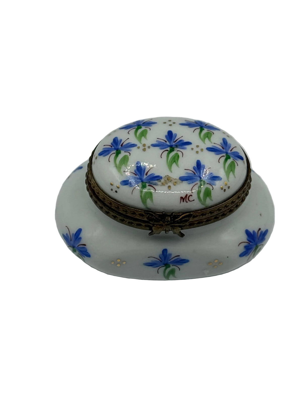 A white porcelain trinket box with a domed lid, decorated with hand-painted blue lilies with green leaves and small gold dots. The box features a brown woven trim around the lid's rim and a small red monogram 'MC' on the lid.