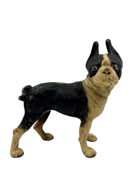 A small painted dog figurine, likely a Boston Terrier, standing on a white surface against a light gray background. The dog is painted black and tan.