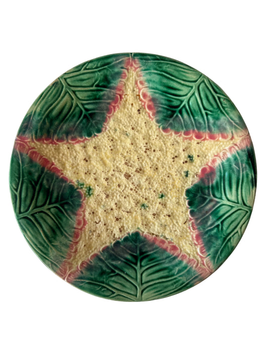 A round ceramic plate featuring a star-shaped center in a textured cream-yellow glaze, surrounded by five leaf-like sections with a green and red glaze pattern, photographed from directly above on a white fabric background.