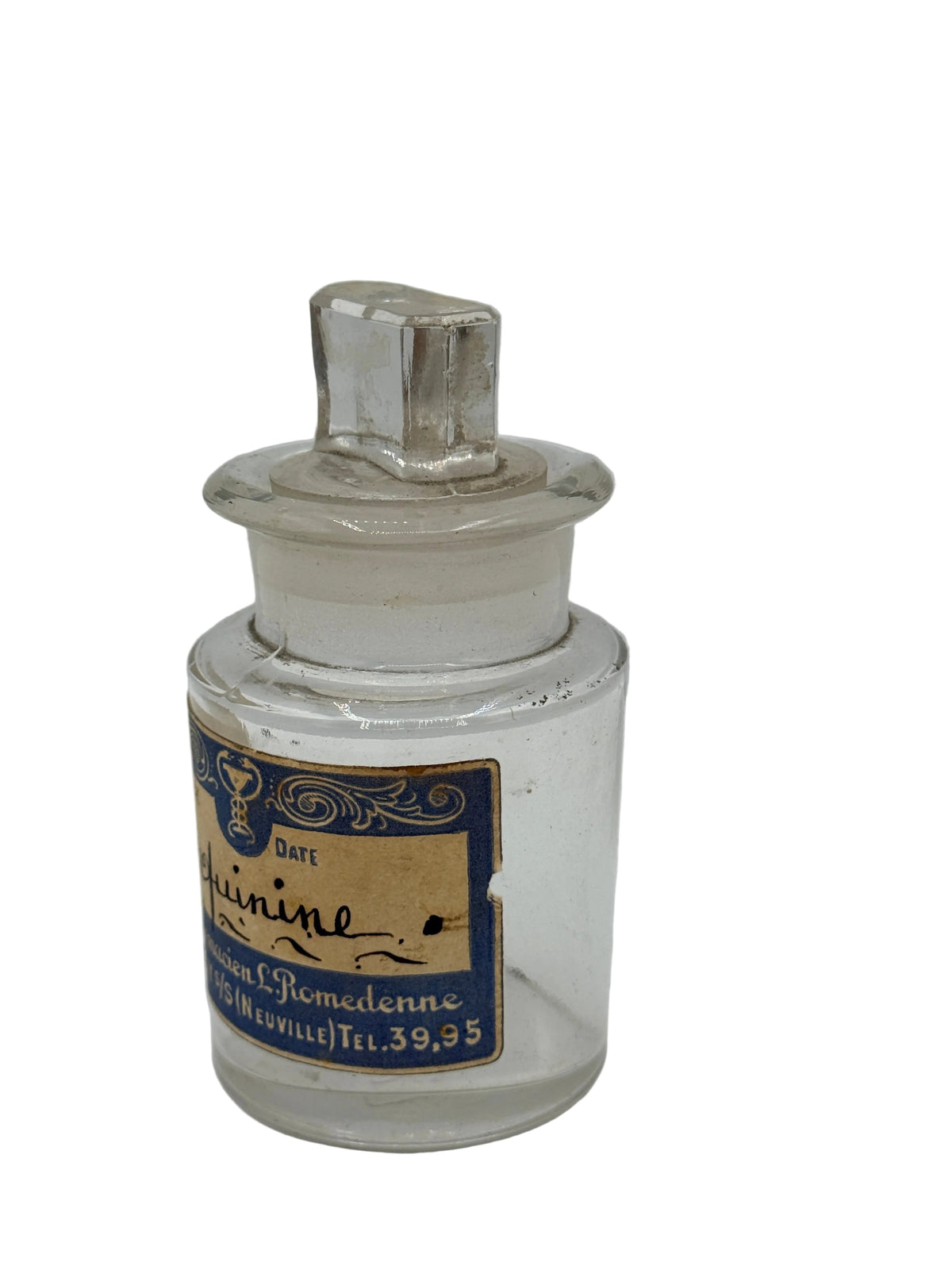A vintage clear glass apothecary bottle with a stopper lid and a blue and beige label, photographed against a neutral light gray background.