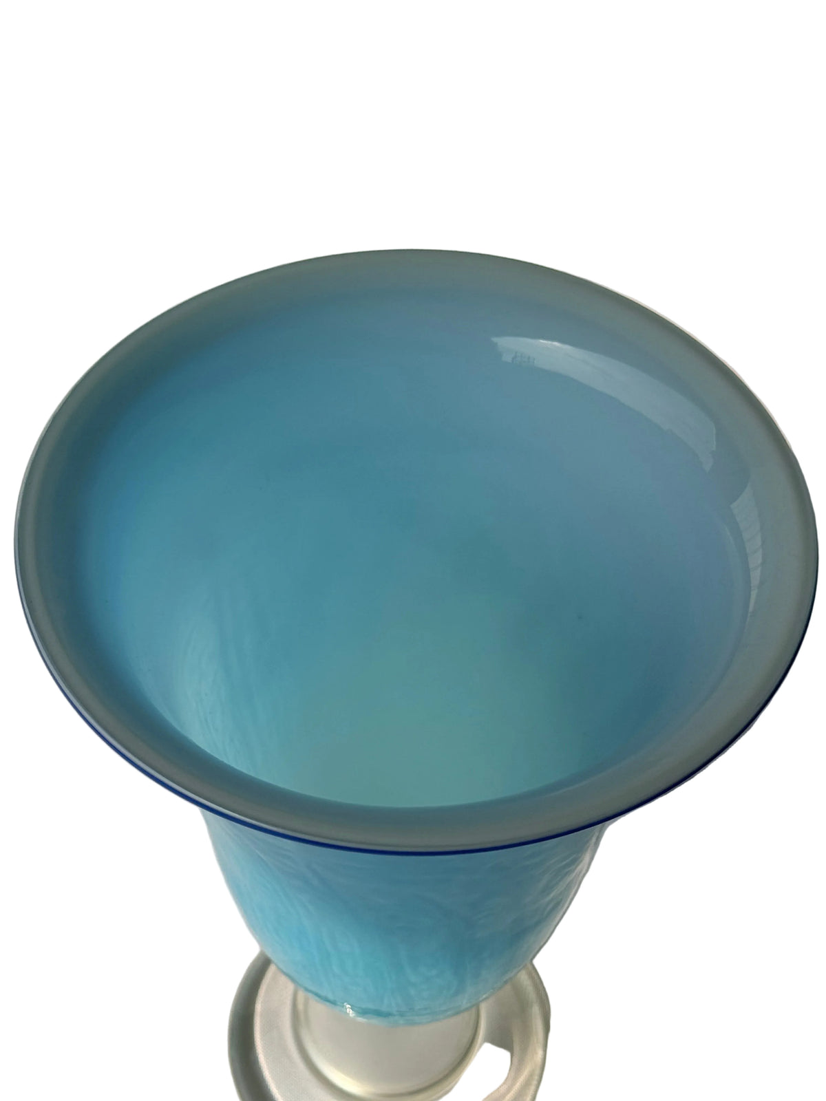 A light blue translucent glass vase with a flared rim and a clear glass base, photographed against a white paneled wall and white surface.