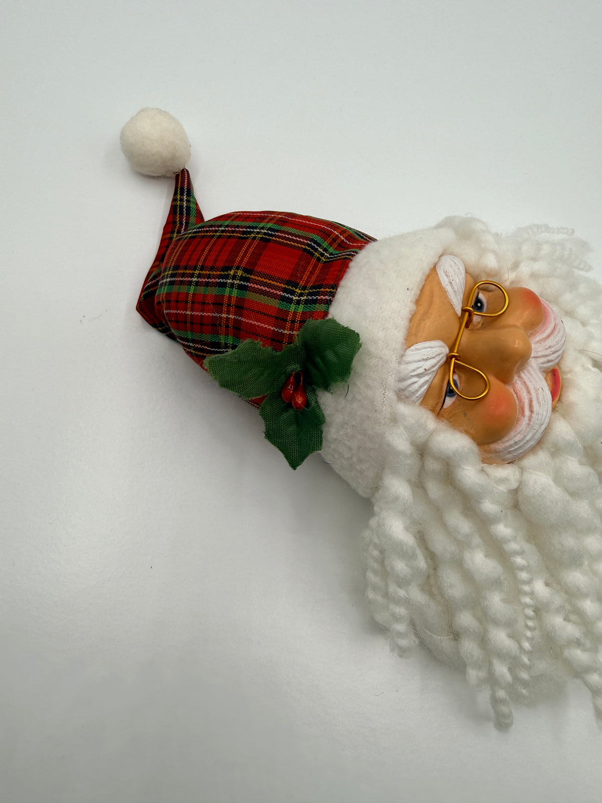 A close-up of a Christmas Santa decoration or plush head with a white textured beard, gold-rimmed glasses, a red tartan Santa hat with green holly accents, and a white pom-pom, photographed on a plain white background.