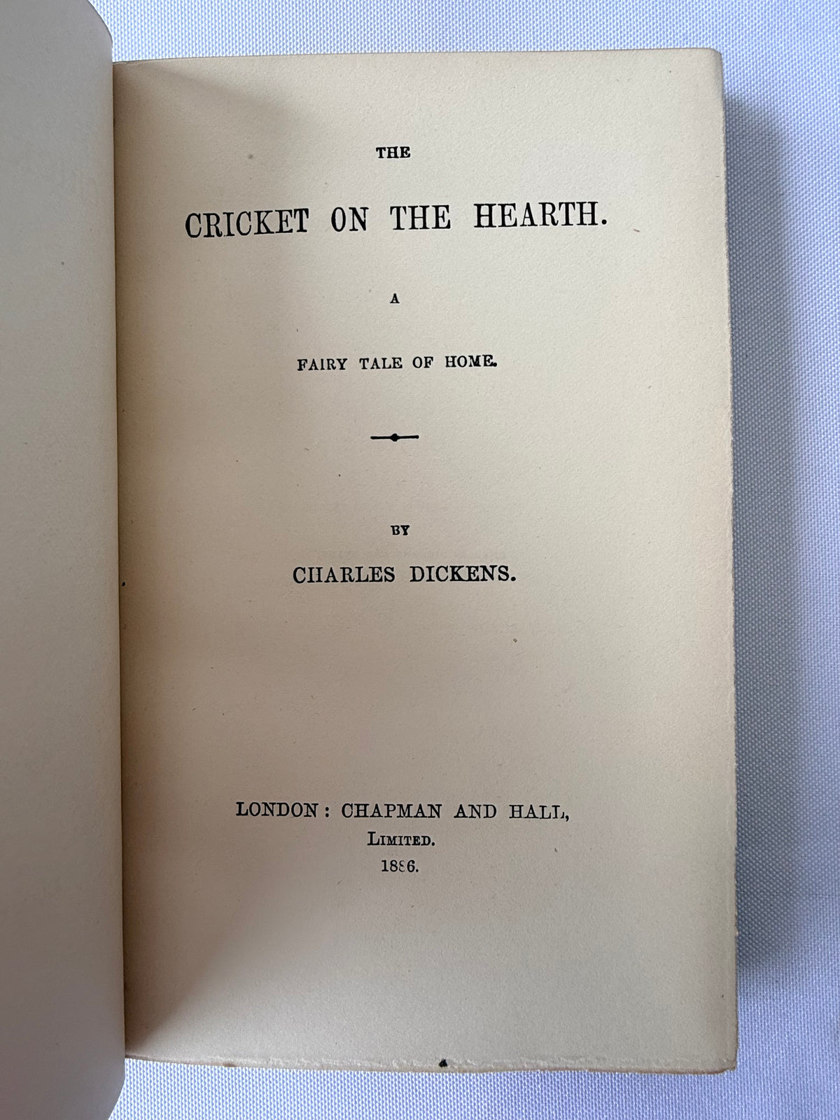 An open vintage book showing the title page of 'The Cricket on the Hearth' by Charles Dickens, printed in black serif text on aged beige paper against a light blue fabric background.