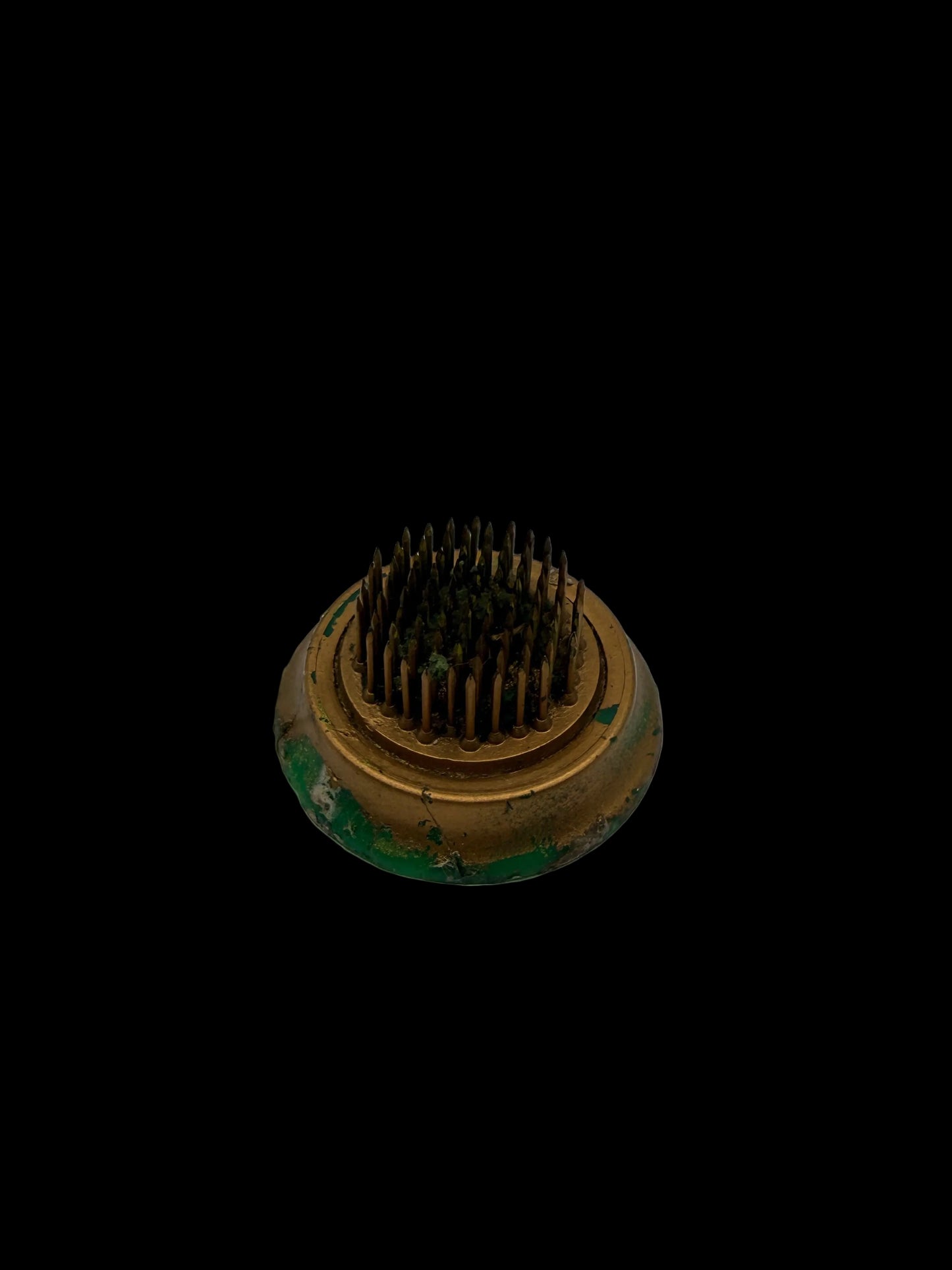 A small round metal grooming brush with dense dark bristles and a tarnished gold-toned base showing green rust or patina, photographed on a plain white surface.