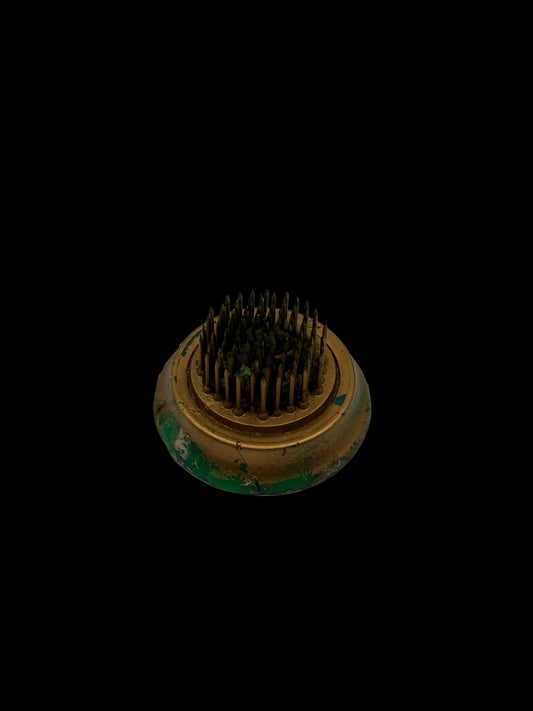 A small round metal grooming brush with dense dark bristles and a tarnished gold-toned base showing green rust or patina, photographed on a plain white surface.