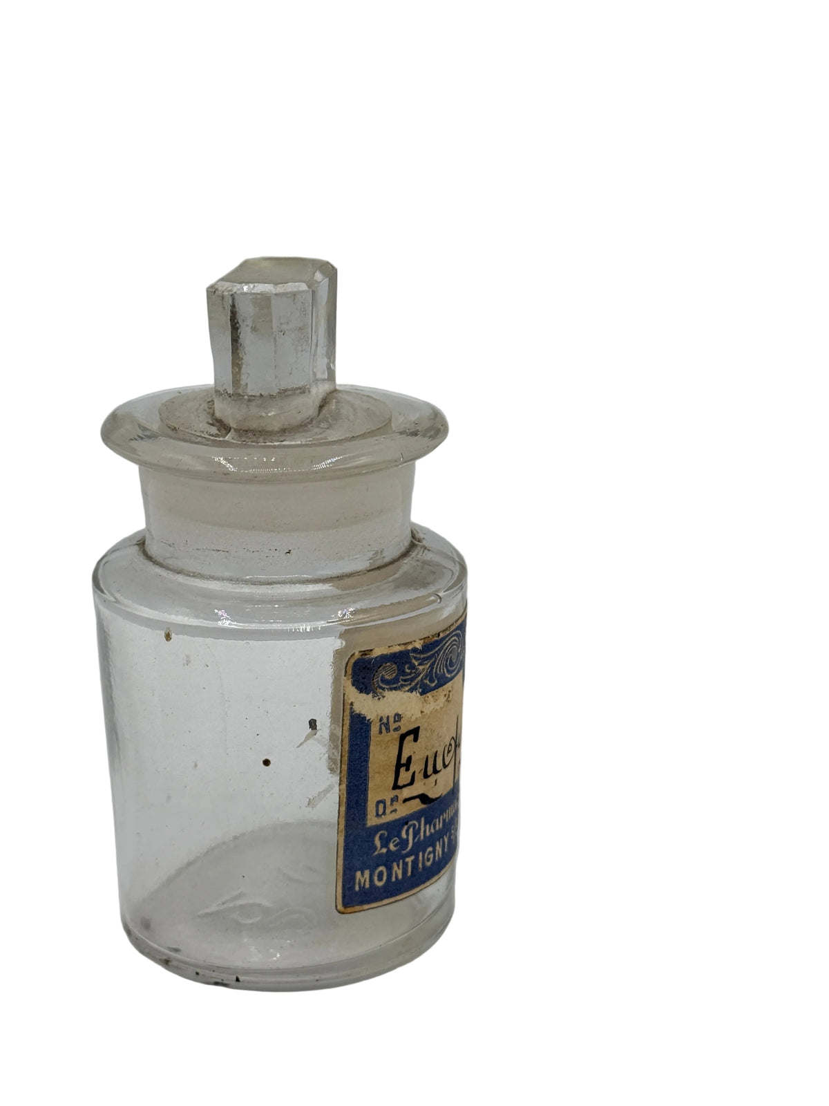 A vintage clear glass apothecary bottle with a stopper lid and a blue and gold label, photographed on a white surface against a light gray background.