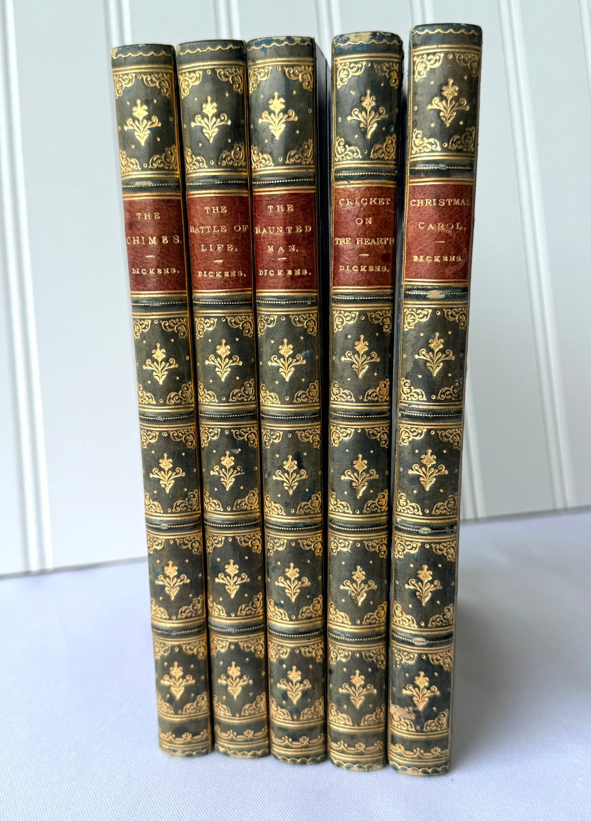 A set of five antique hardcover books standing upright, featuring dark green leather spines with ornate gold foil detailing and red cloth bands with gold lettering.