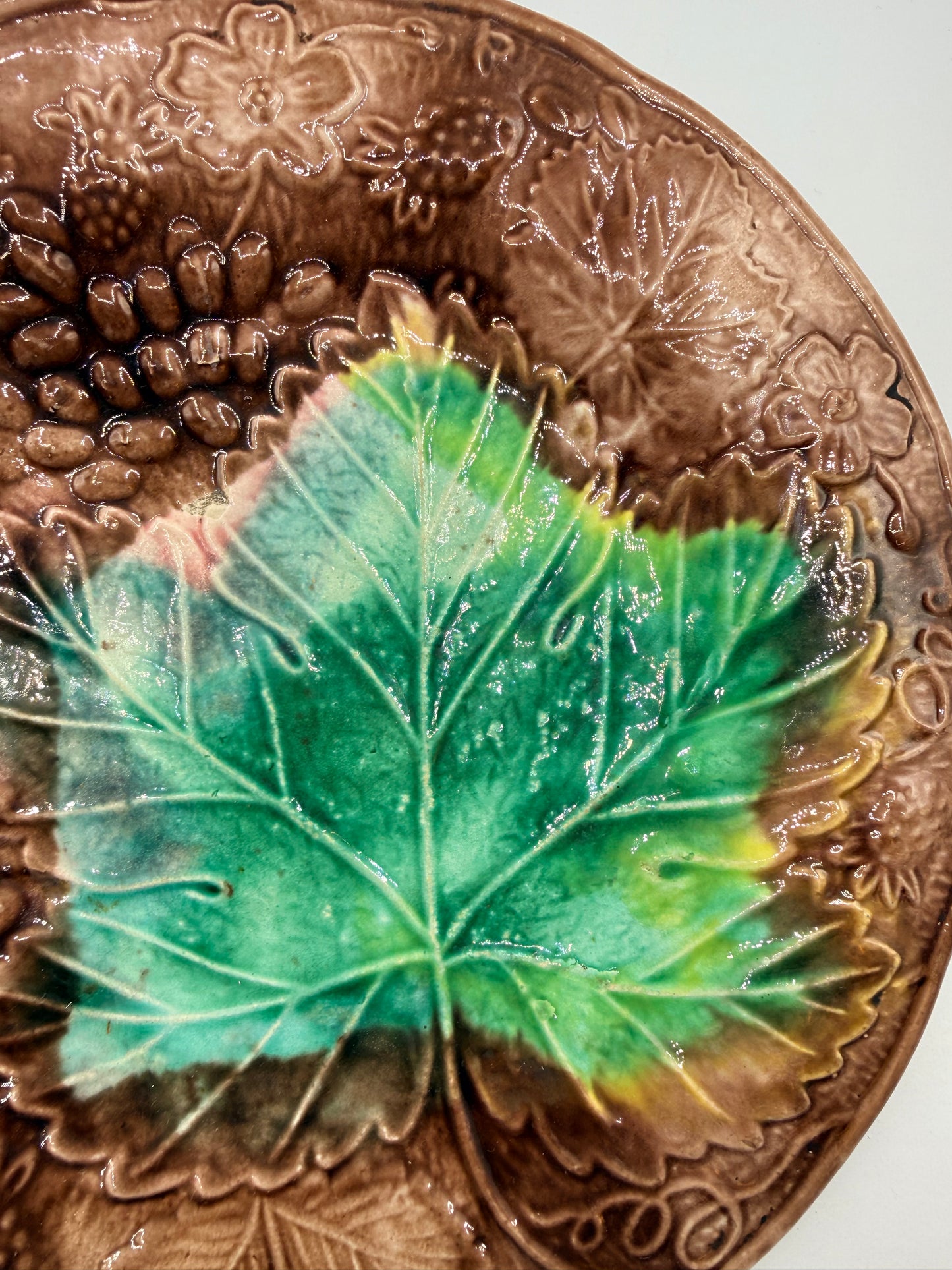 A close-up of a brown embossed ceramic plate featuring a glossy green leaf with yellow and pink accents, set against a light gray background.