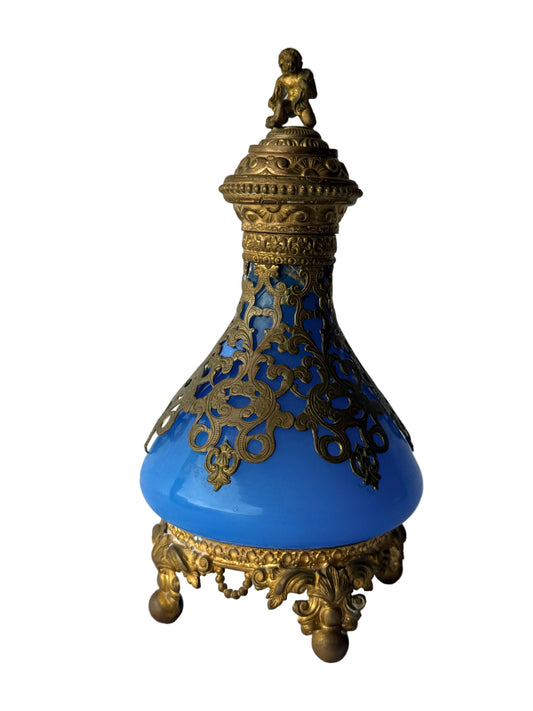 An ornate blue glass bottle with intricate gold filigree detailing, a matching gold lid topped with a small figurine, and a decorative gold base, set against a white paneled background.