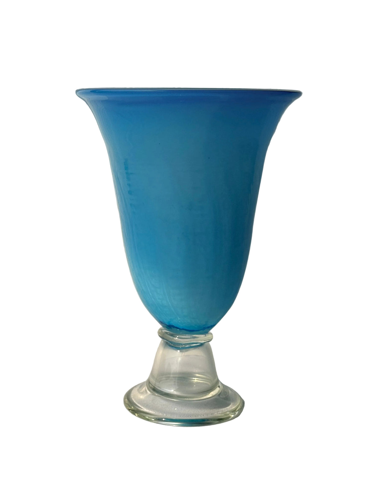 A cobalt blue glass vase with a flared rim and a clear glass pedestal base, set against a white paneled wall with soft sunlight and shadows.