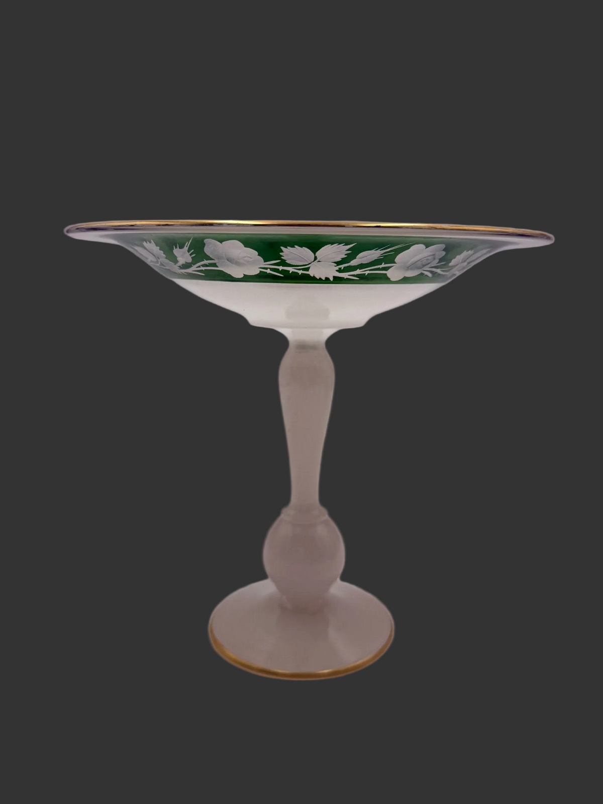 A vintage champagne coupe glass featuring a pale pinkish-beige stem, a white interior, and a green exterior decorated with white floral motifs and gold trim.