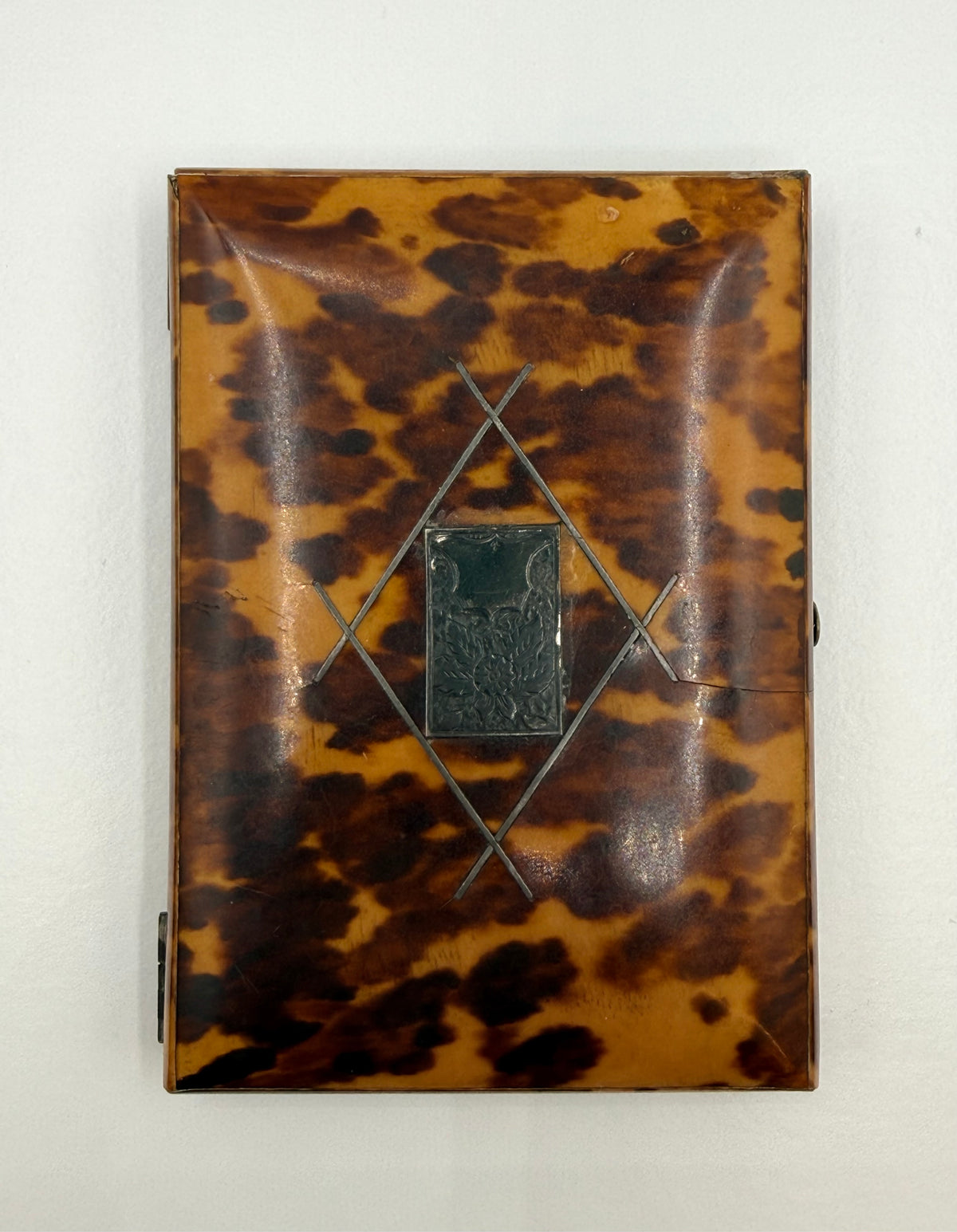 A vintage rectangular case with a glossy tortoiseshell pattern in amber-brown and black, featuring a central dark metal plaque and geometric metal accents, shown against a plain off-white background.