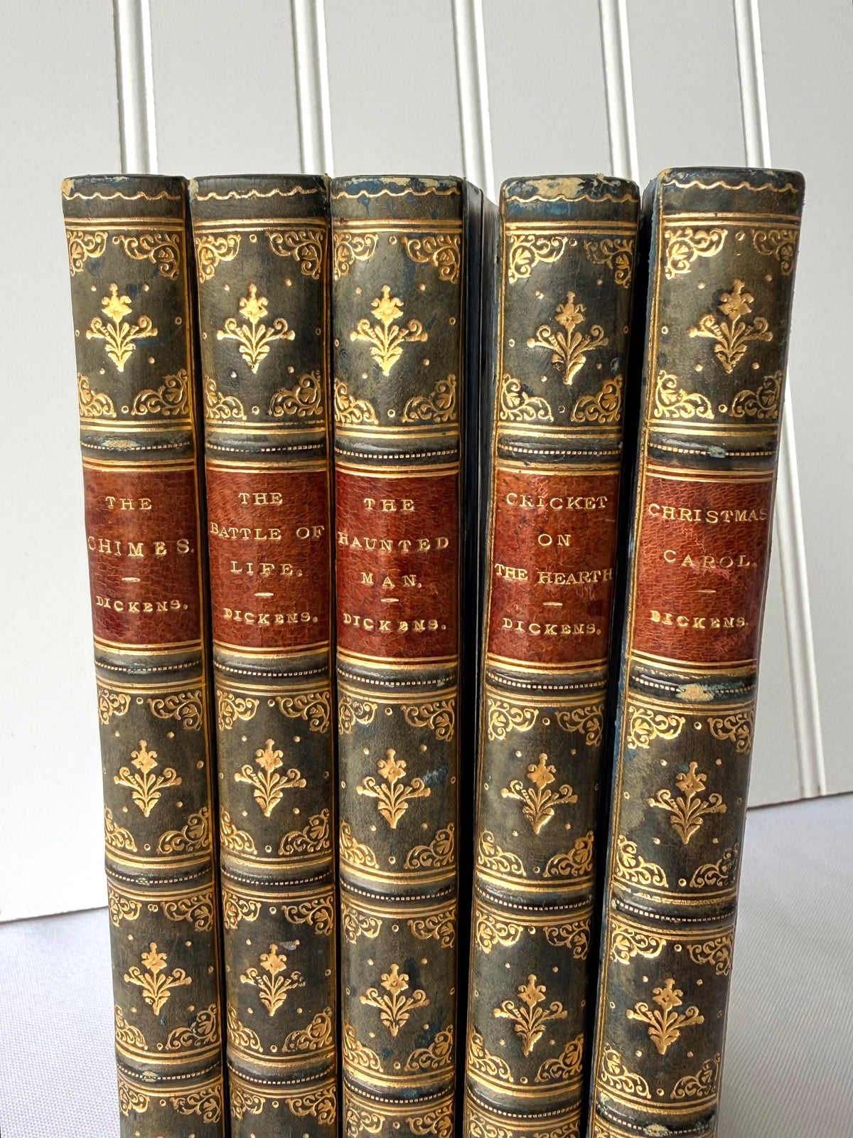 Five antique hardcover books standing upright, showing their decorative dark green and gold leather-bound spines with red title bands. Visible titles include The Chimes, The Battles of Life, Cricket on the Heath, Christmas Carol, and Dickens.