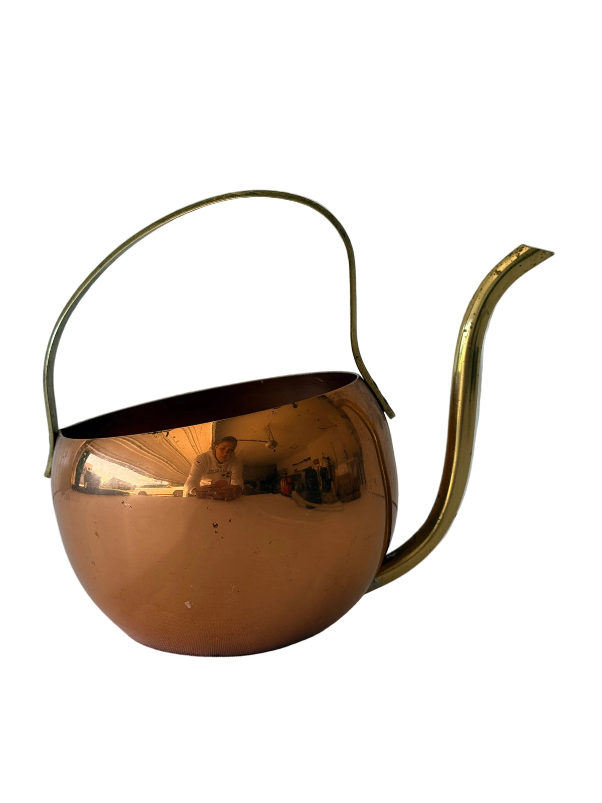 A polished copper teapot with a brass loop handle and a curved spout, shown on a light surface against a white paneled background.