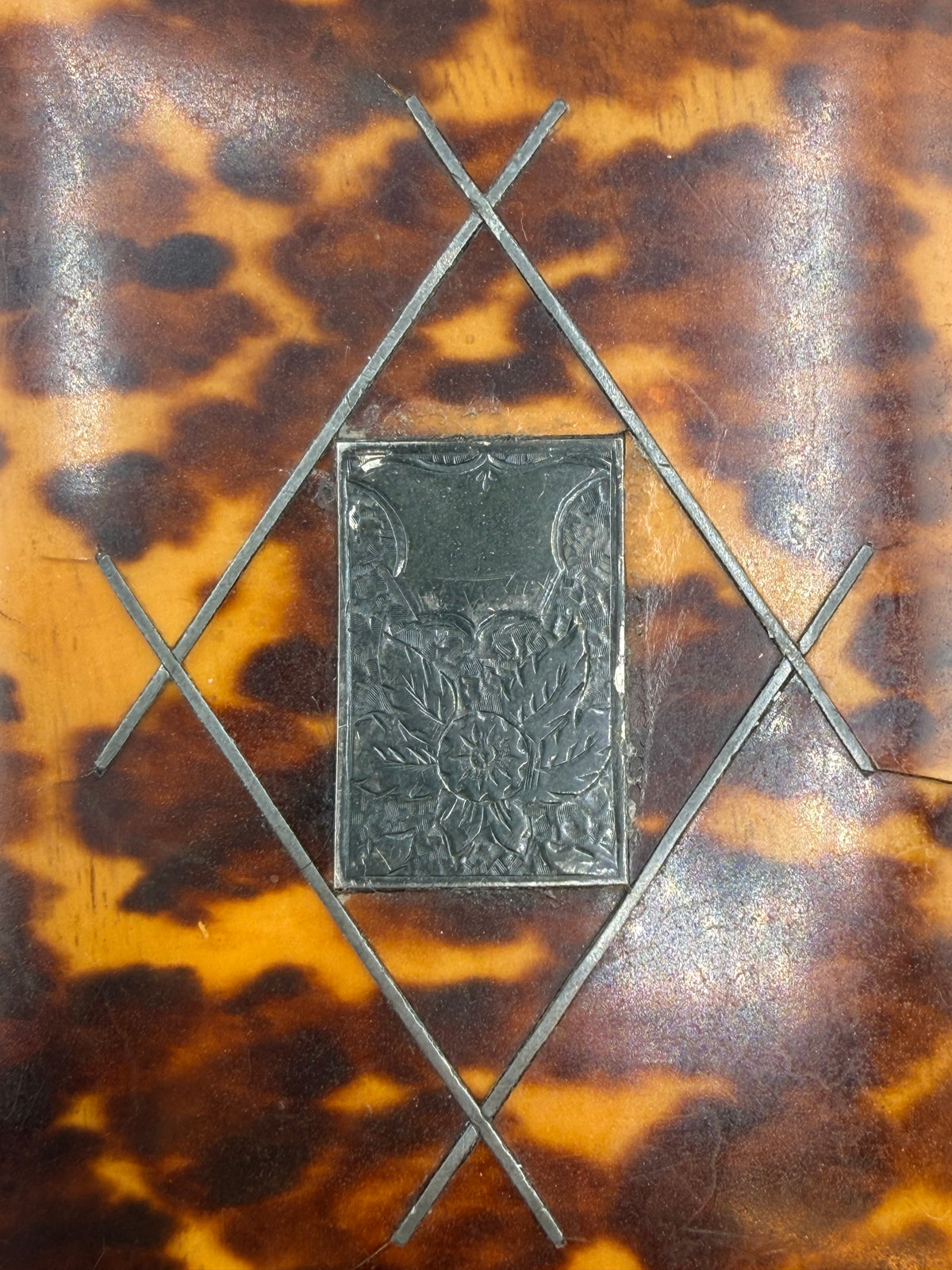Close-up of a vintage tortoiseshell box or case featuring a brown and amber mottled pattern, with a central dark metal plaque and geometric metal frame details.