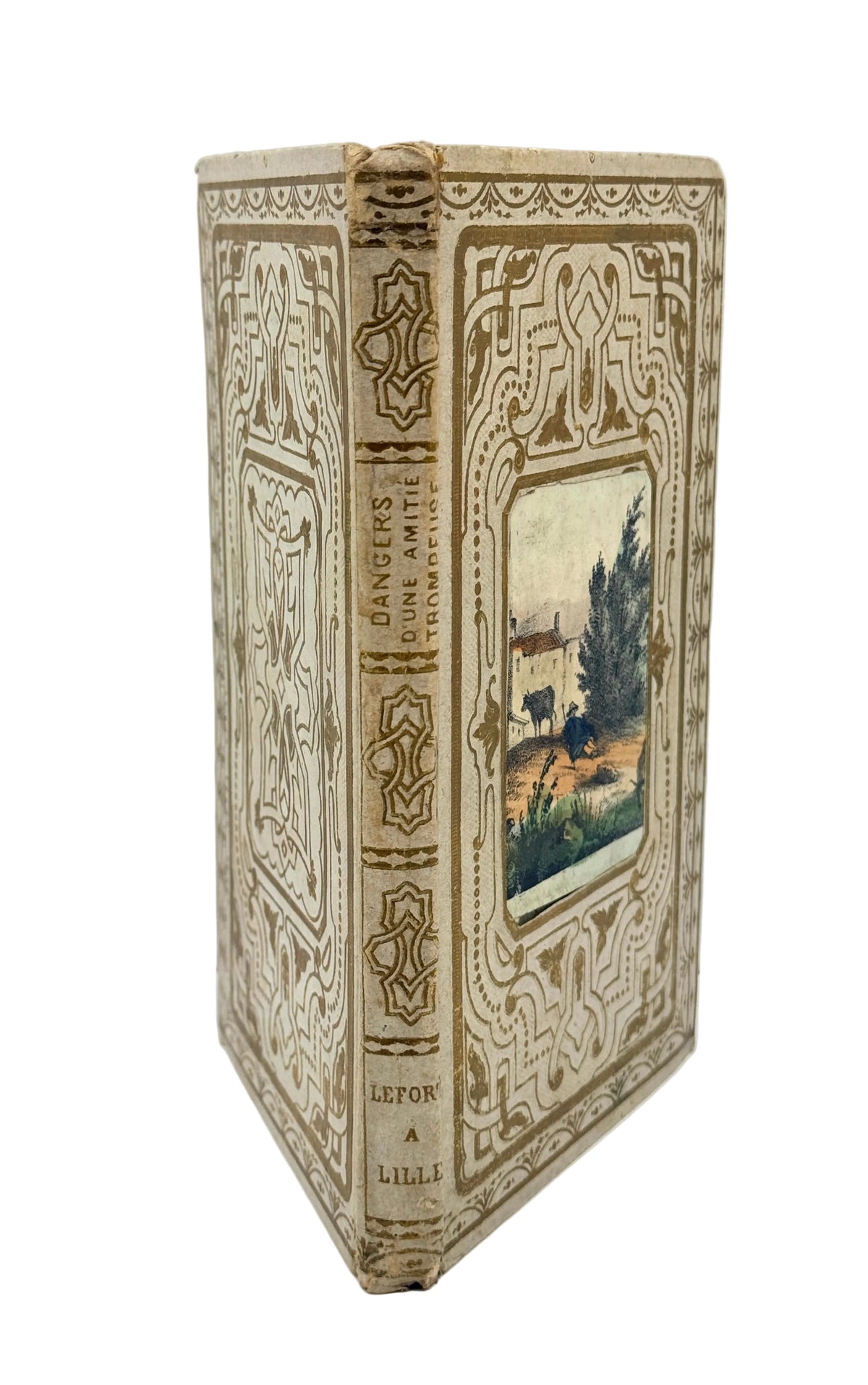 An antique hardcover book standing upright, showing its ornate cream-colored leather-bound spine and decorated front cover with a small central landscape illustration.