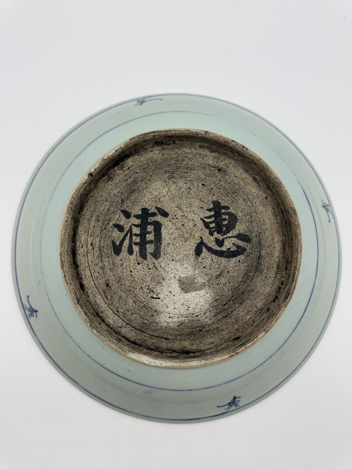 Top-down view of a ceramic plate or dish base with a light blue-green glaze, blue painted motifs near the rim, and black calligraphic characters in the center of the dark brown interior.