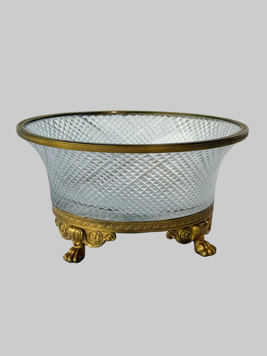 An ornate vintage-style glass bowl with a clear cut diamond pattern, framed by a polished gold-tone metal rim and matching gold feet, set against a white paneled background.