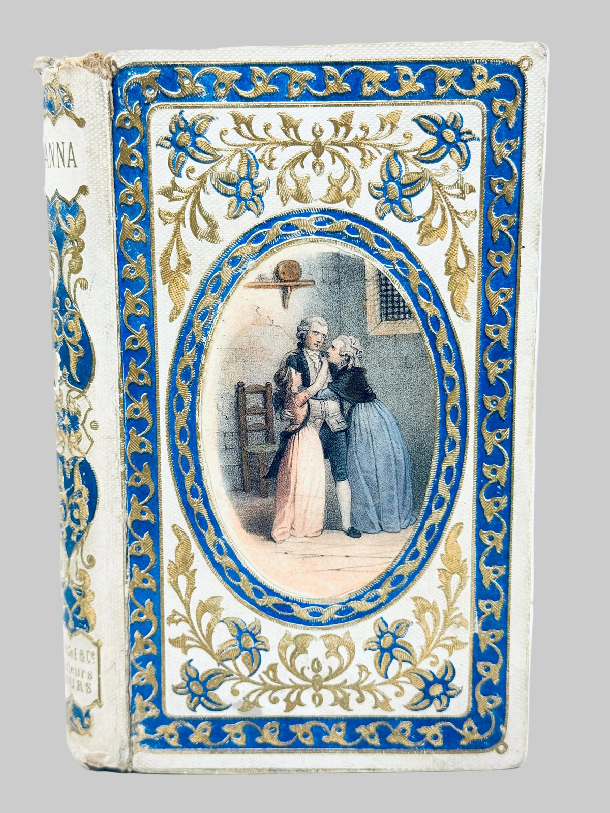 An antique hardcover book titled 'Anna' with ornate blue and gold floral detailing on a cream-colored cover, featuring a central oval illustration of two women.
