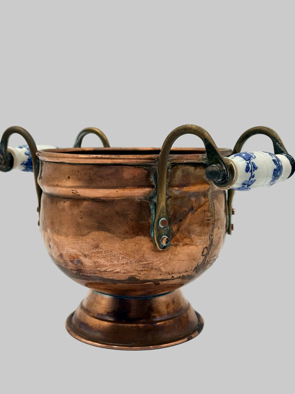 A vintage copper bowl with a pedestal base, featuring four curved metal handles and white ceramic grips decorated with blue floral patterns, set against a neutral gray background.