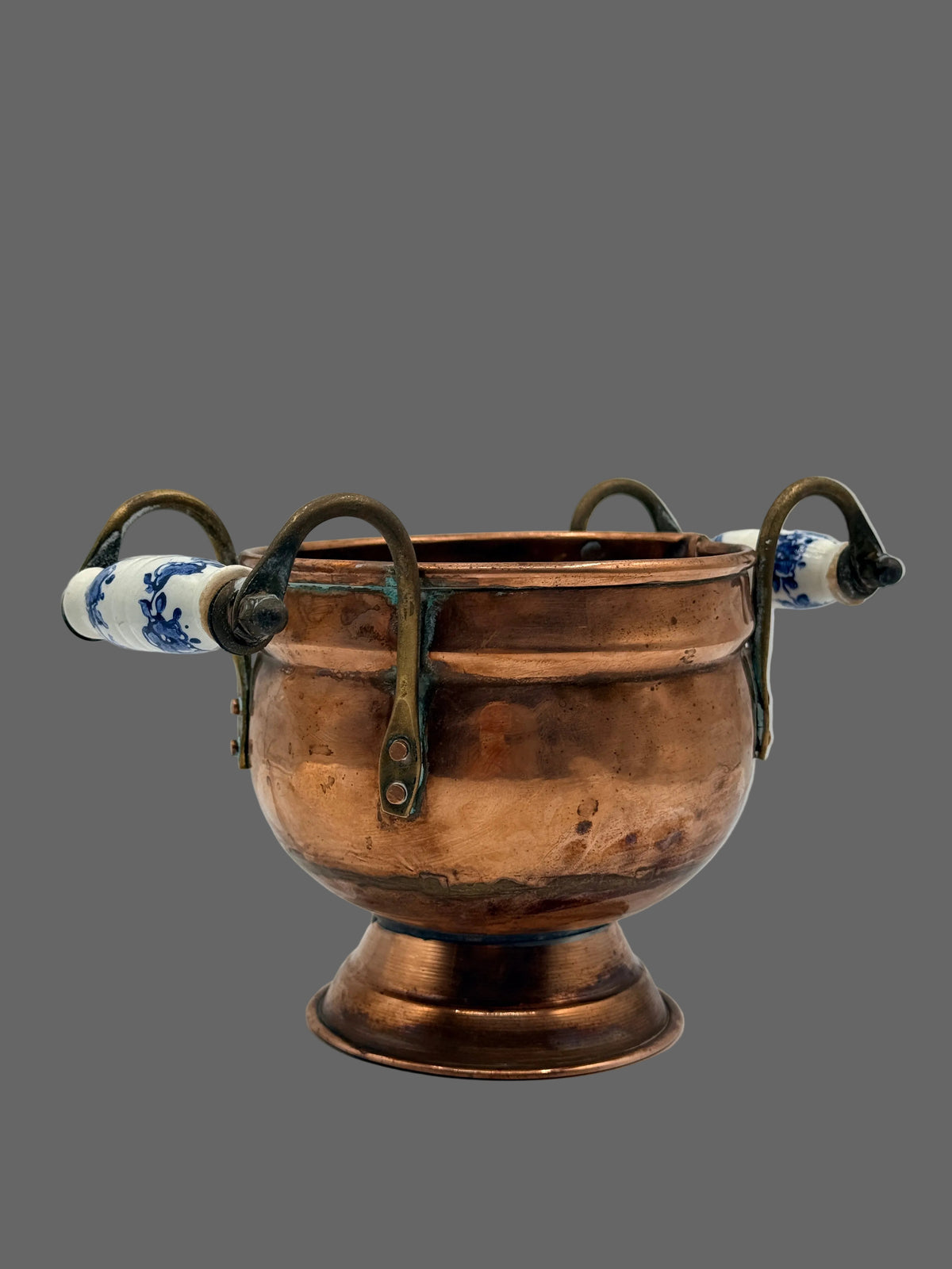 A vintage copper chalice or cup with four loop handles and blue-and-white ceramic accents on the ends, photographed on a neutral light gray background.