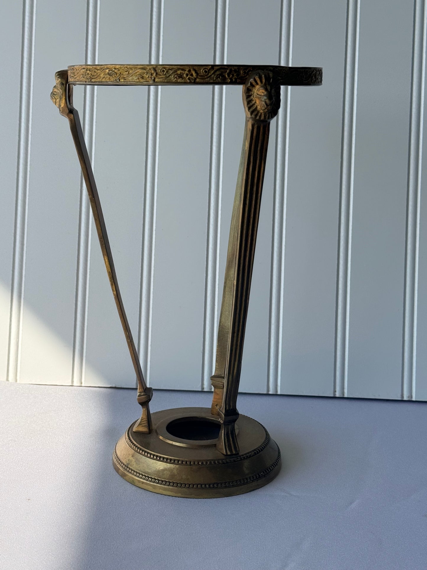 An ornate antique-style brass candlestick holder with a decorative top bar, two vertical stem supports, and a round base, set against a white paneled wall.
