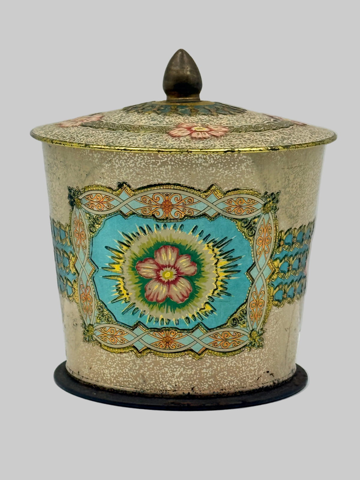 An antique decorative lidded tin or canister with a textured beige body, featuring a central turquoise blue floral medallion with pink and yellow details, surrounded by gold and green ornamental borders. The top has a dark metal knob, and the base sits on a round black stand, photographed against a neutral light gray background.
