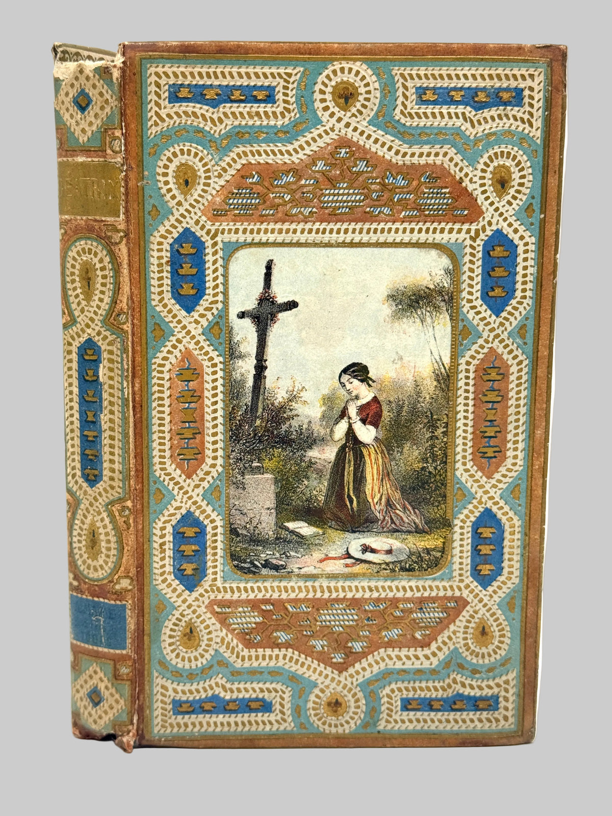 An antique hardcover book with ornate decorative cover and spine, featuring a central painted illustration of a woman kneeling before a cross in a garden.