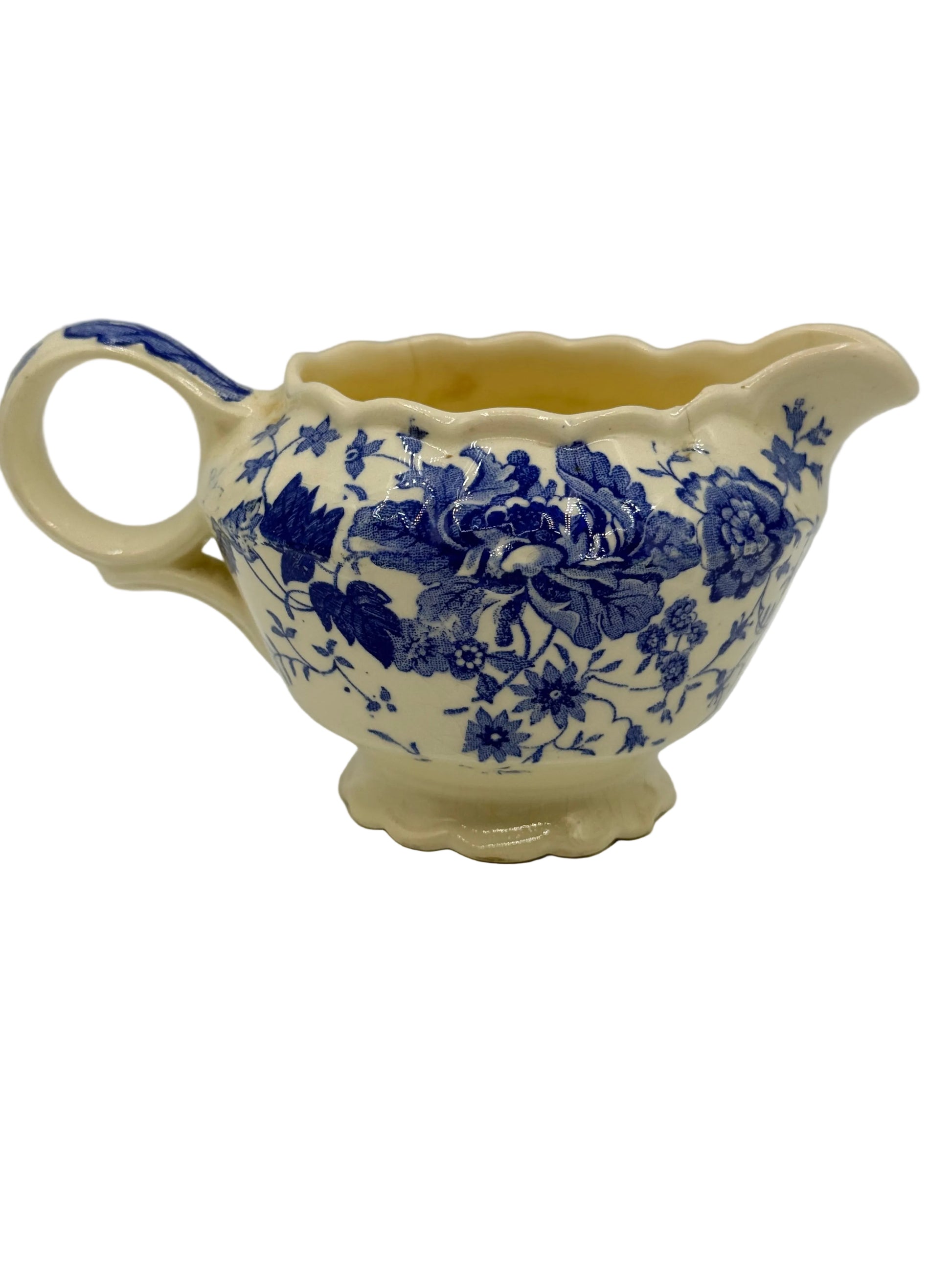 A vintage cream-colored ceramic creamer or sauceboat with a blue floral pattern, featuring a scalloped rim and base, set against a neutral gray background.