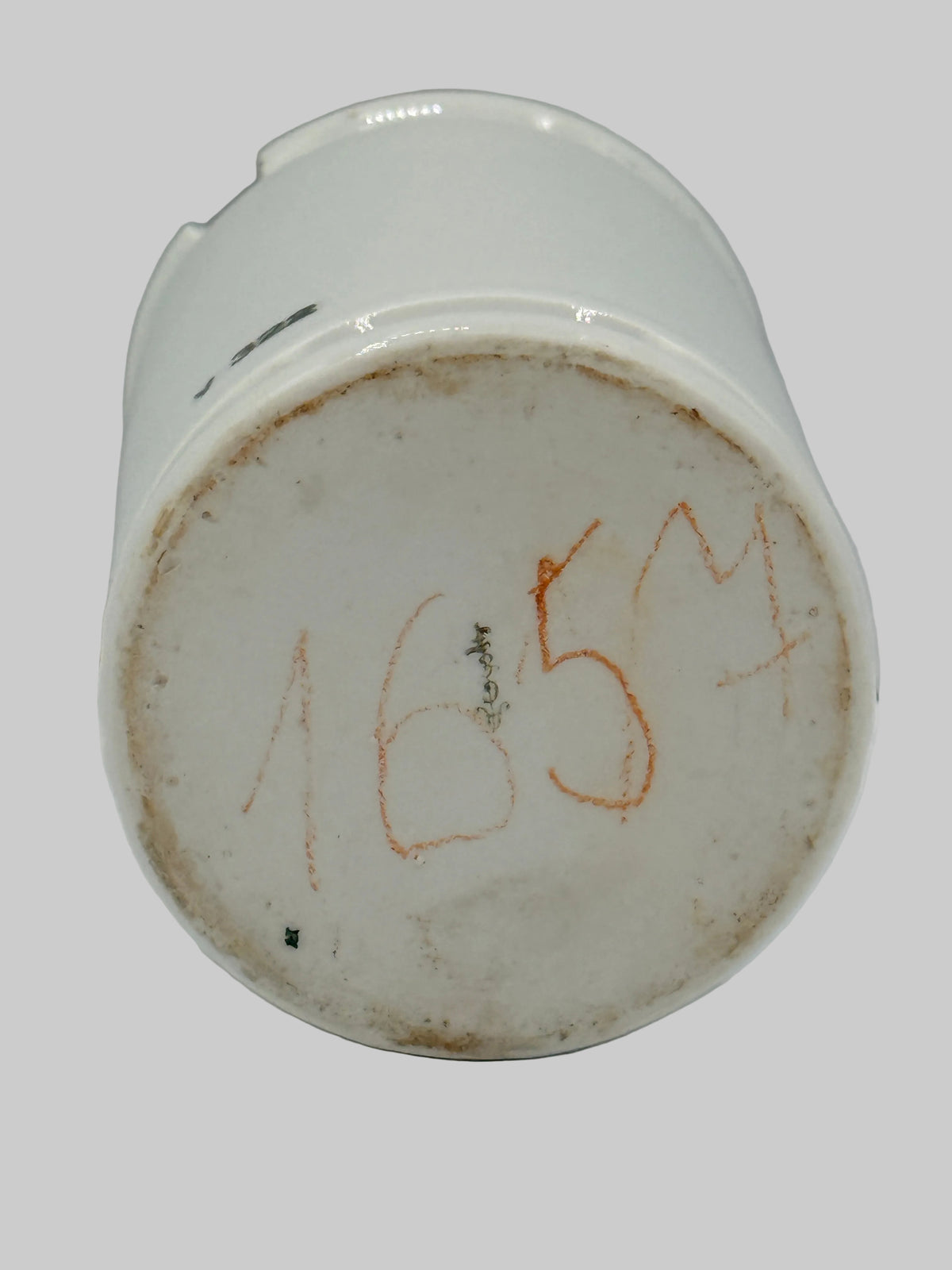 The bottom base of a white ceramic cup or small vase, showing a round off-white glazed surface with handwritten orange numbers '1654' and some dark markings, photographed against a plain light background.
