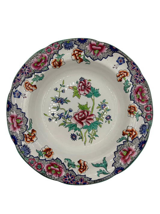 A round ceramic plate with a white base, decorated with a colorful floral pattern featuring pink roses, green leaves, and blue and orange blossoms, set against a plain light gray background.