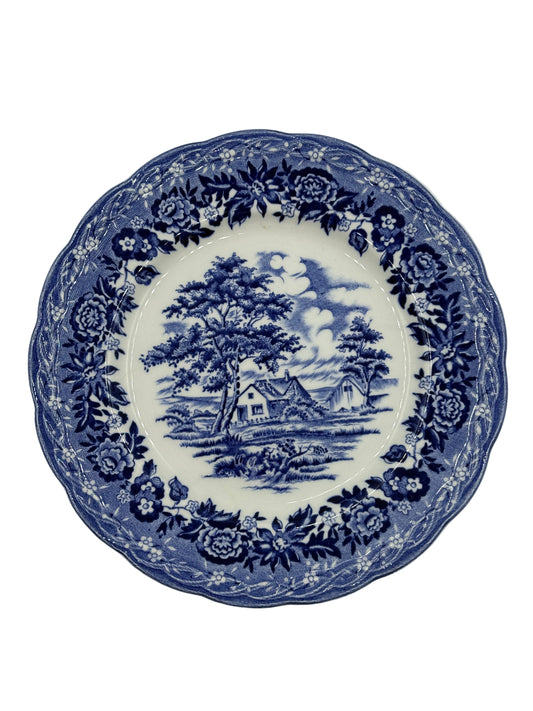 A blue and white ceramic plate featuring a classic transfer design with a central landscape scene of trees and cottages under a moonlit sky, surrounded by a floral border.