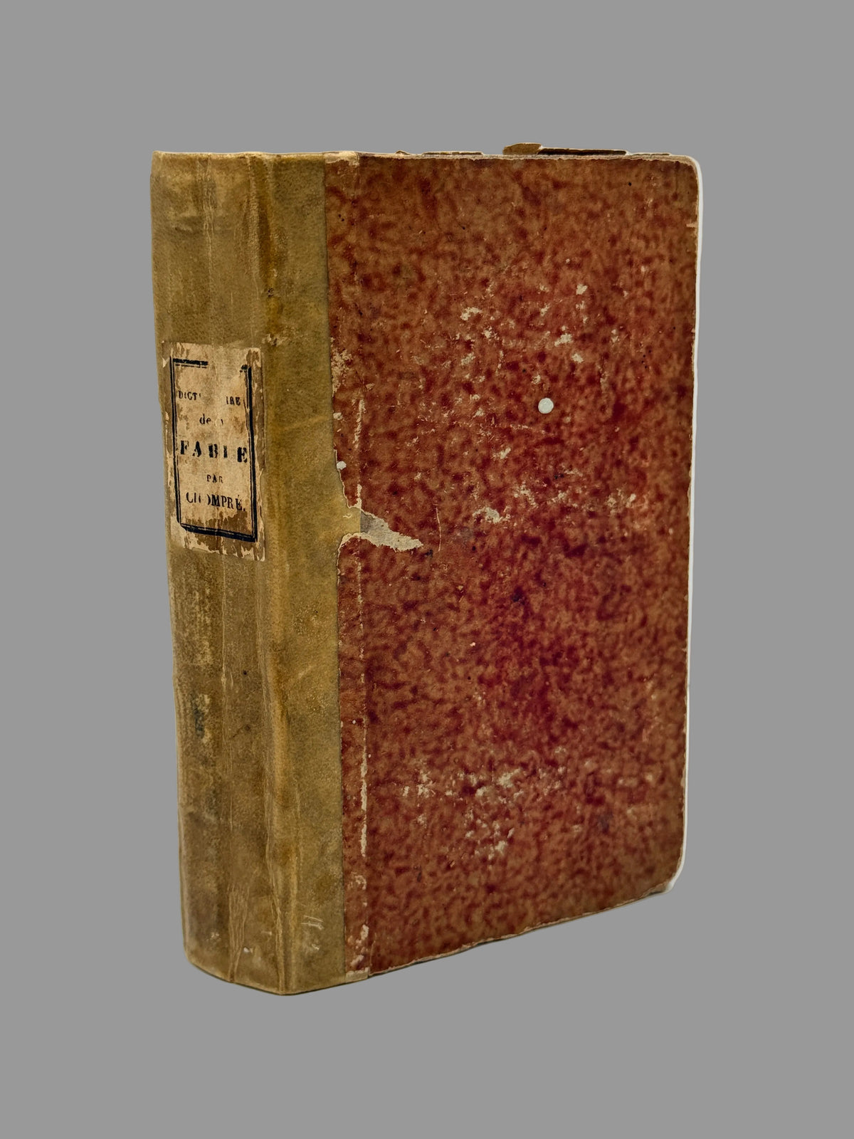 An antique hardcover book with a textured reddish-brown front cover and a tan spine, standing upright on a white surface against a light gray background.