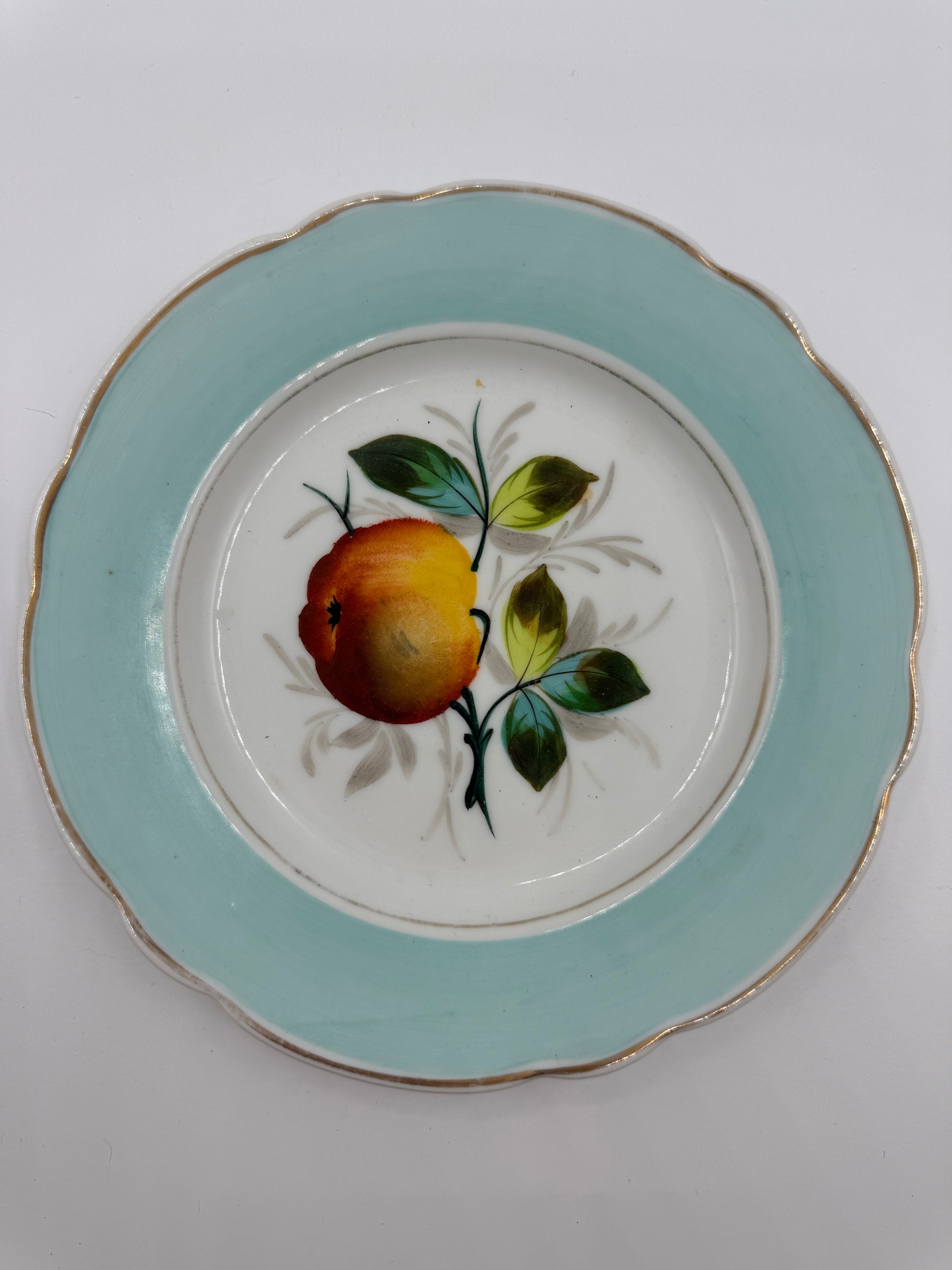 A vintage ceramic plate with a scalloped rim, featuring a light blue outer border, a white center, and a hand-painted fruit motif with green leaves and gold trim.