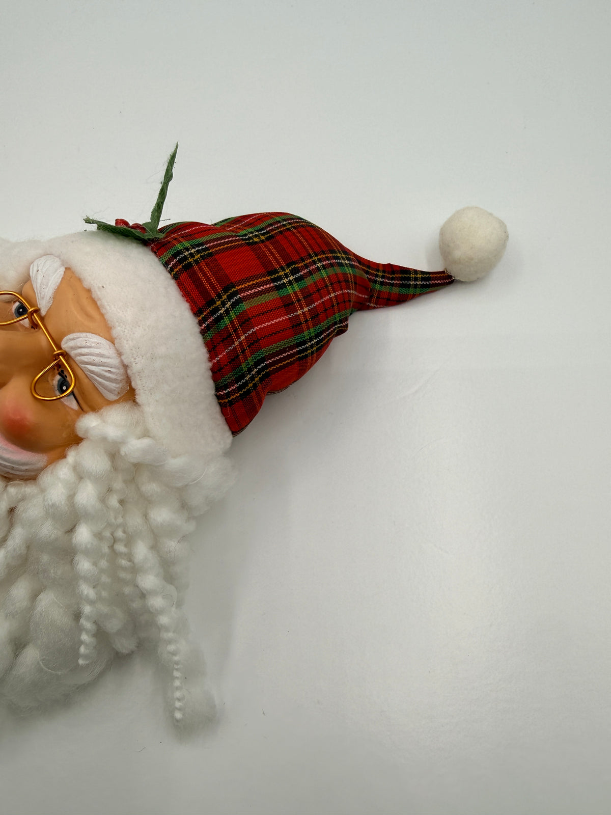 A close-up of a Santa Claus decorative head with a red tartan plaid hat, white fluffy beard, white faux fur trim, and gold-rimmed glasses on a plain white background.