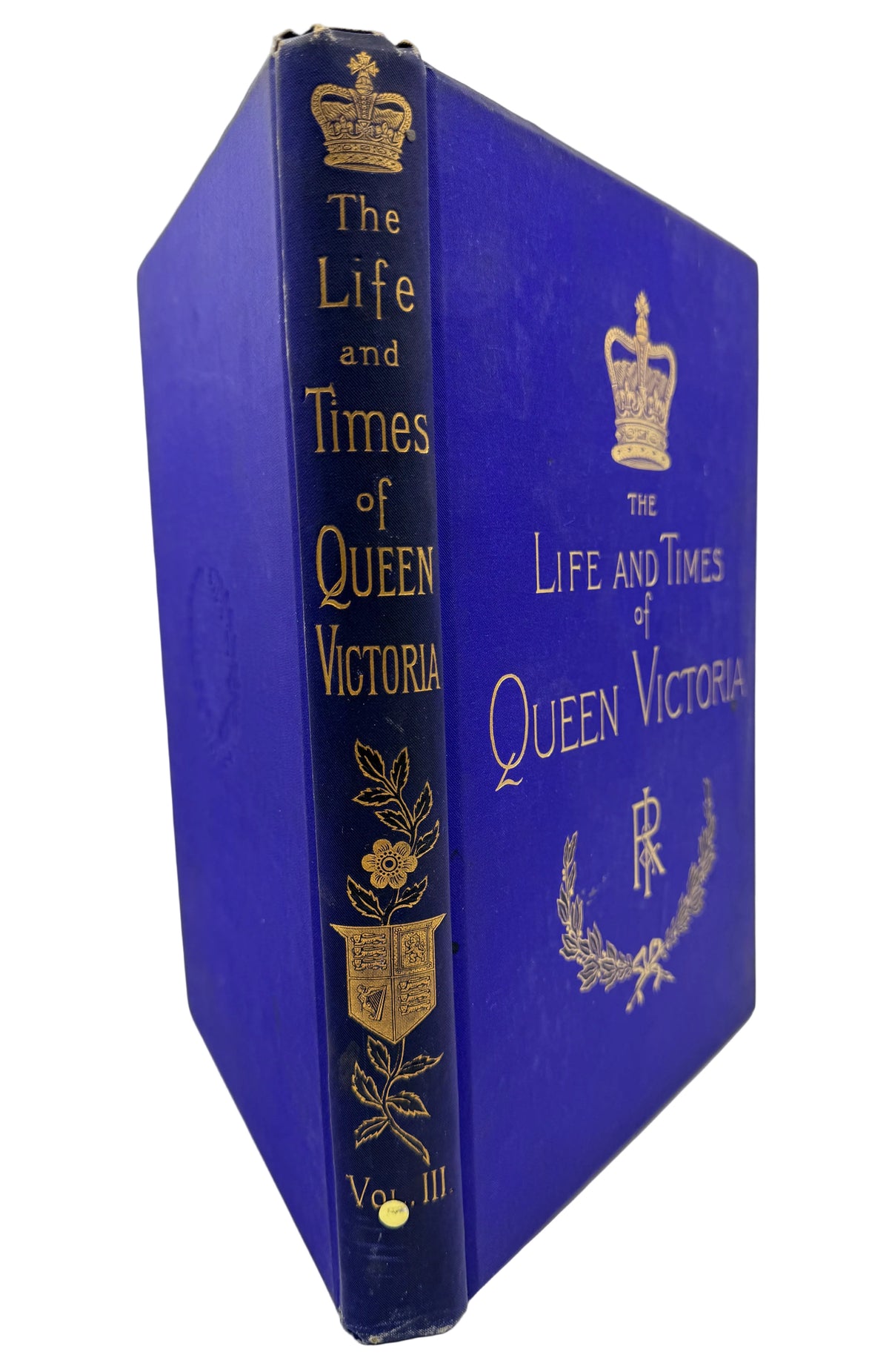 A vintage hardcover book titled 'The Life and Times of Queen Victoria' shown standing upright at an angle. The book has a deep blue cloth cover with gold foil detailing, including a crown, title text, floral motifs, and an initial monogram on the front cover.