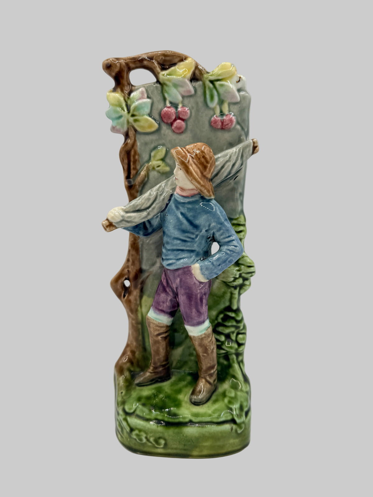 A vintage ceramic vase featuring a sculpted figure of a man carrying a large basket on his shoulder, set against a painted vineyard backdrop with cherries and green foliage.