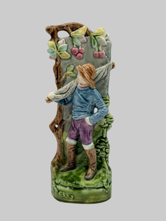 A vintage ceramic vase featuring a sculpted figure of a man carrying a large basket on his shoulder, set against a painted vineyard backdrop with cherries and green foliage.