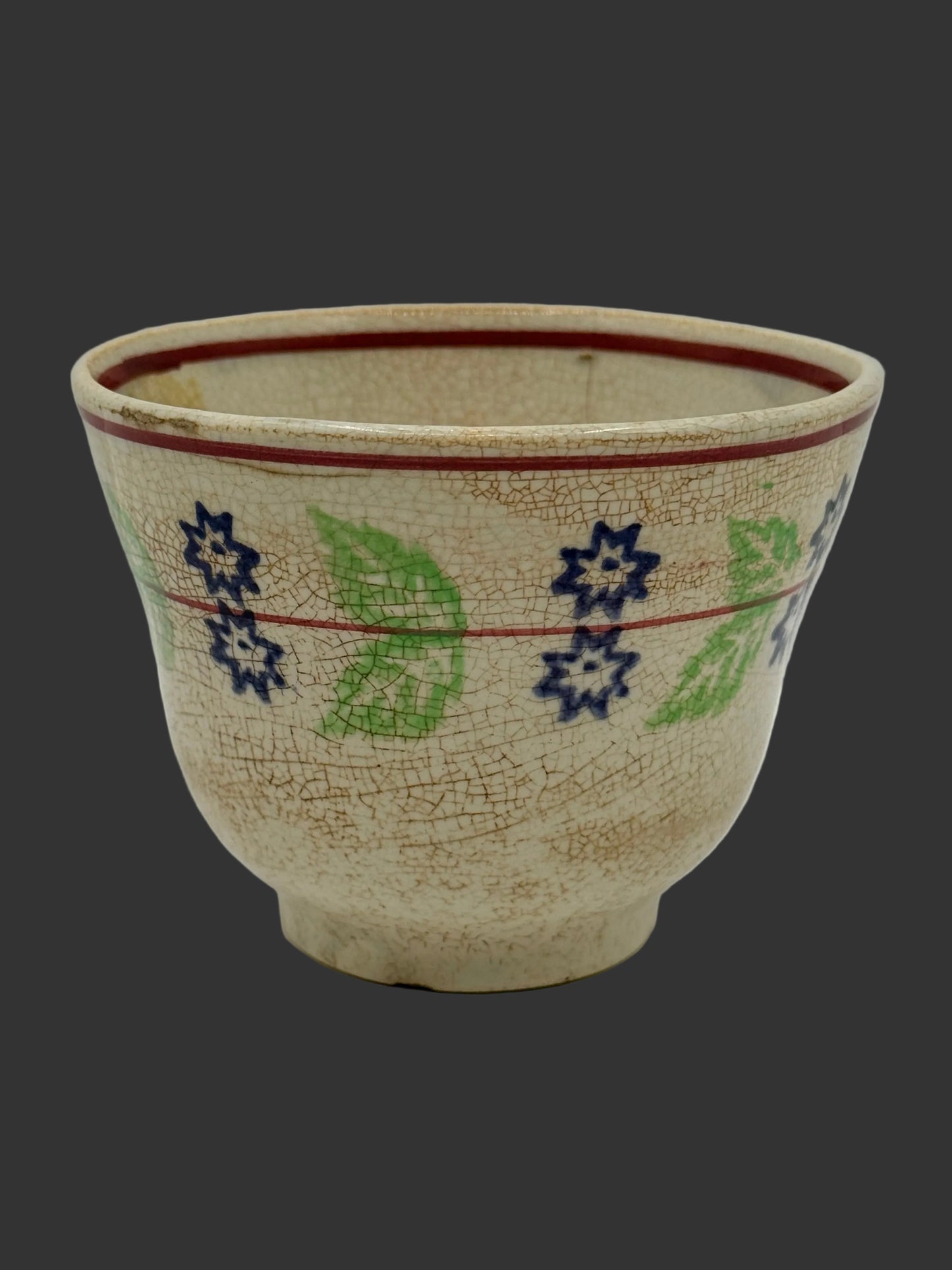 A vintage ceramic bowl with a crackled beige glaze, decorated with blue and green floral and leaf patterns and red painted lines, set against a plain light gray background.