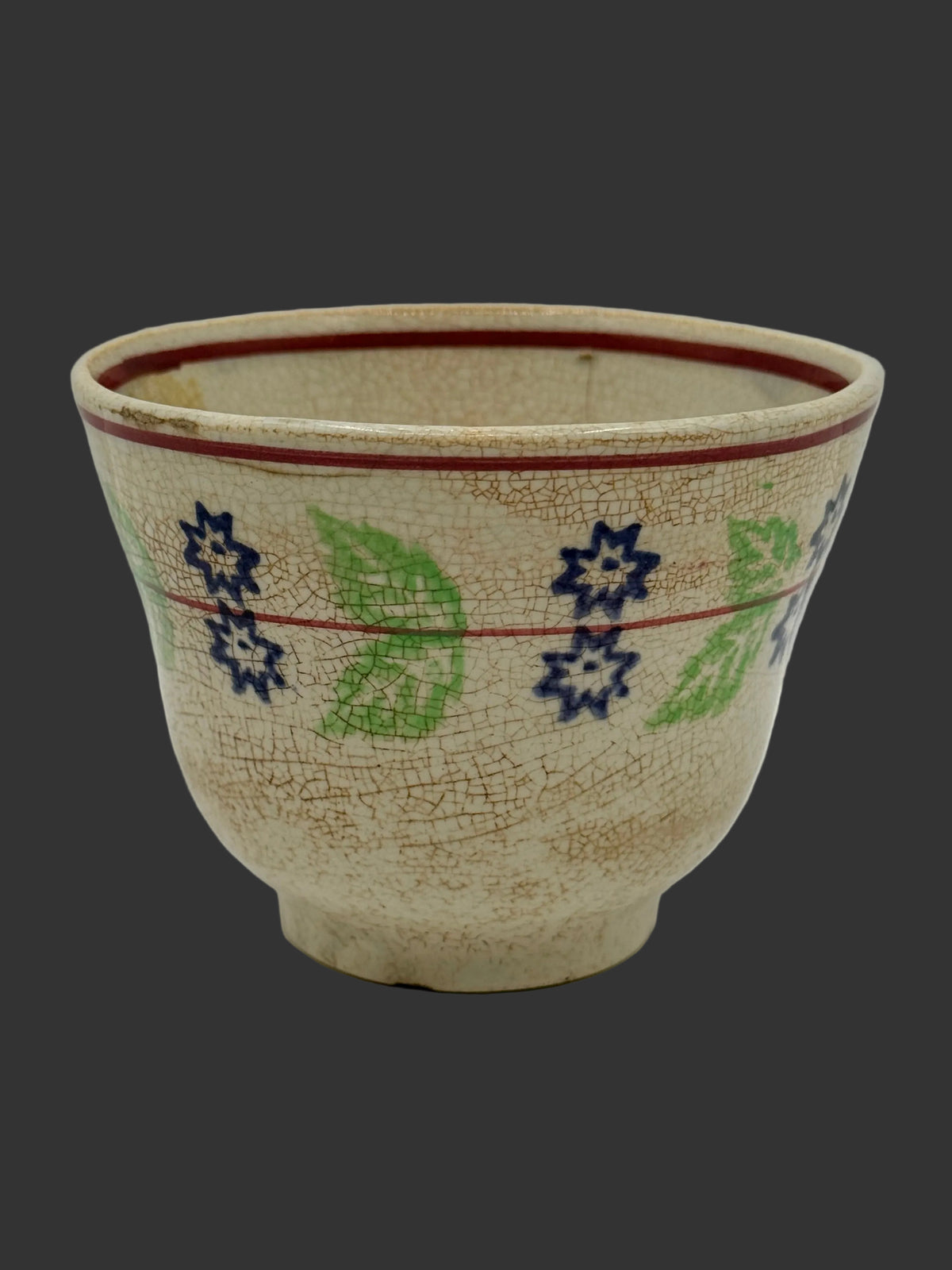A vintage ceramic bowl with a crackled beige glaze, decorated with blue and green floral and leaf patterns and red painted lines, set against a plain light gray background.