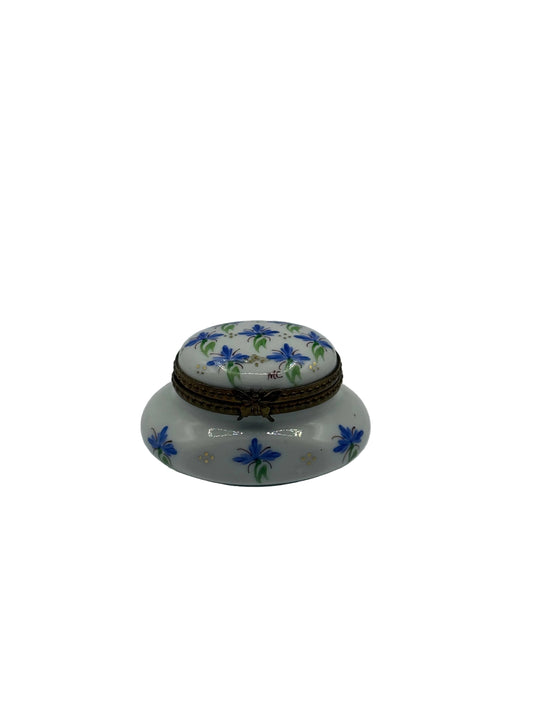 A small white ceramic lidded box decorated with blue flowers and green leaves, featuring a brown braided cord closure, set against a plain light gray background.