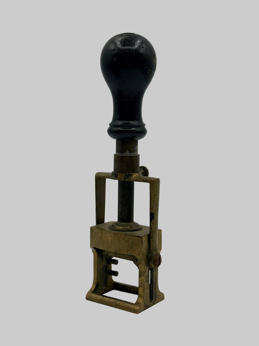 A vintage manual stamp with a dark, rounded wooden handle and a weathered brass or bronze metal frame base, photographed on a neutral light gray background.