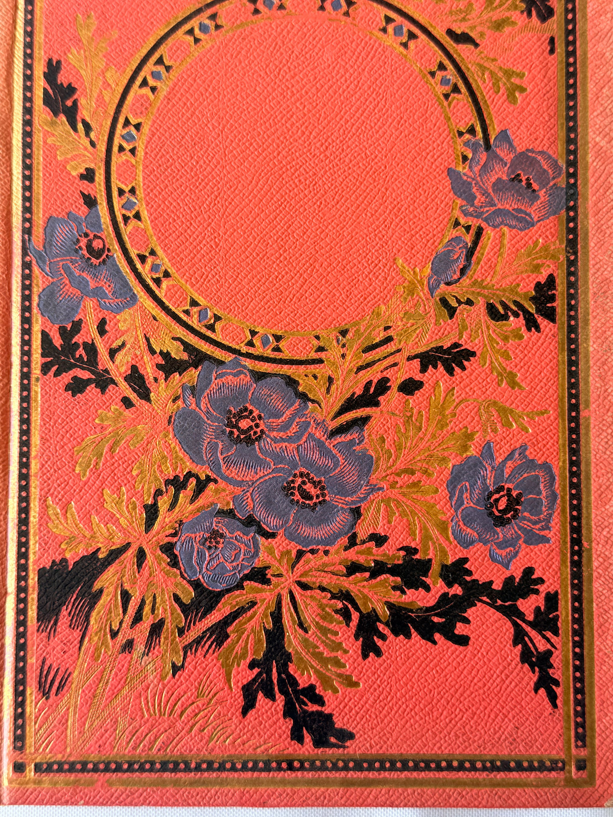 A close-up of a decorative textile or printed surface featuring a coral-pink background with a large circular motif, blue floral blossoms, and black-and-gold leafy stems.